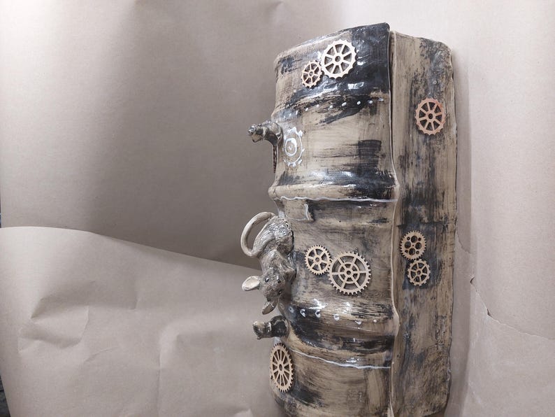 May include: A cylindrical ceramic sculpture in a steampunk style. It has a textured, aged appearance with black and beige tones. Wooden gear accents and a small animal sculpture are incorporated into the design.