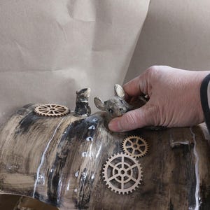 May include: A ceramic sculpture with a wood-grain effect, featuring small animal figurines and wooden gear accents. The sculpture has a glossy finish and is being held by a hand.