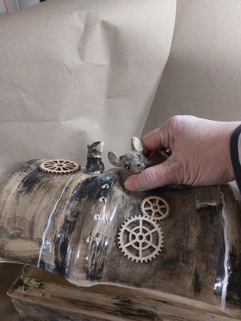 May include: A ceramic sculpture with a wood-grain effect, featuring small animal figurines and wooden gear accents. The sculpture has a glossy finish and is being held by a hand.