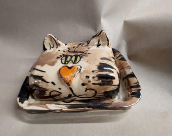 May include: A ceramic butter dish shaped like a cat head with a glossy finish. The piece features a cream-colored base with brown and black painted tabby stripes, yellow eyes, and a prominent orange heart-shaped nose on the lid.