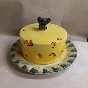 Può includere: Un burriera in ceramica gialla a forma di formaggio con una piccola statuetta di creatura nera sul coperchio. Il coperchio presenta rientranze arancioni che ricordano i buchi del formaggio svizzero. La base ha un motivo radiale verde e crema.