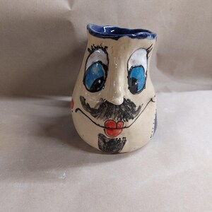 Könnte beinhalten: Ein Keramikbecher mit einem handbemalten Gesicht, das große blaue Augen, einen schwarzen Schnurrbart, einen kleinen Ziegenbart und rote Lippen zeigt. Der Becher hat eine cremefarbene Basis, einen dunkelblauen Innenrand und eine unregelmäßige Form.