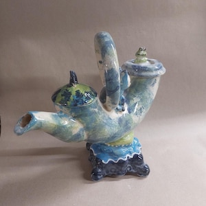 Op de afbeelding: Een grillige keramische theepot in blauw, groen en wit. De theepot heeft een gebogen tuit, een grote handgreep en een deksel met een kikkerfiguur. De basis is donkerblauw met een geschulpte rand. Een uniek decoratief item.