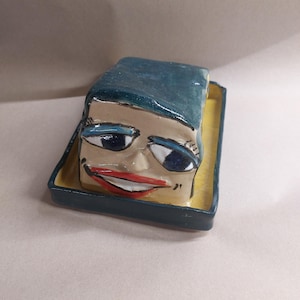 Puede incluir: Una escultura de cerámica caprichosa de una cara con ojos azules y una boca roja, coronada con una cabeza de color turquesa. La cara se asienta sobre una bandeja amarilla con un borde azul oscuro. Una pieza decorativa única.