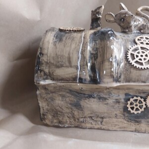 May include: A handcrafted, steampunk-style box with a distressed, textured finish in shades of brown and grey. The box is adorned with wooden cogs and features a small mouse figurine and a small, upright animal figurine on top. A small wooden disc is also on top.