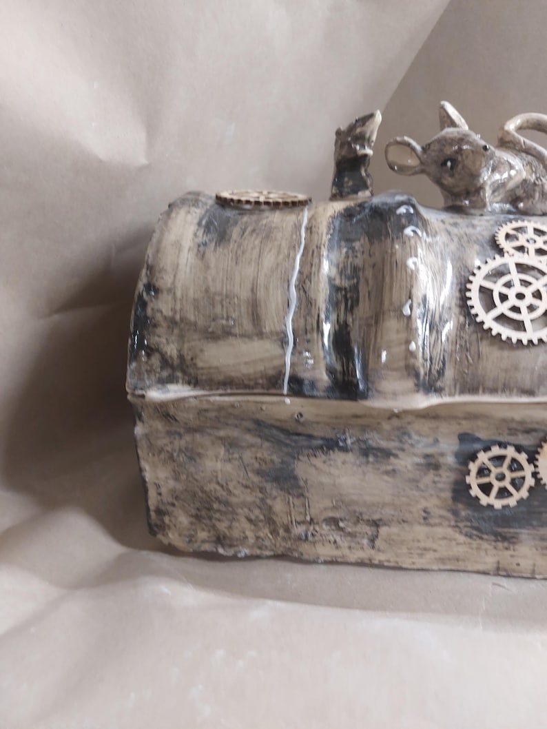 May include: A handcrafted, steampunk-style box with a distressed, textured finish in shades of brown and grey. The box is adorned with wooden cogs and features a small mouse figurine and a small, upright animal figurine on top. A small wooden disc is also on top.