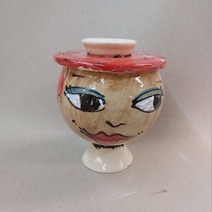 Peut inclure: Un pot en céramique avec un couvercle, peint pour ressembler à un visage avec une peau brune, des yeux bleus et des lèvres rouges. Le pot a une base blanche et un couvercle rouge.