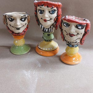 May include: Three ceramic egg cups with painted faces. Each cup has a different facial expression and features a yellow, orange, and green base.