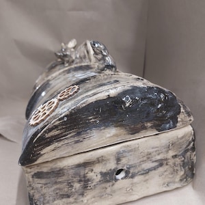 May include: A handmade ceramic box with a lid. The box is mainly off-white with black and grey accents, and a wood-grain texture. Decorative wooden gears are affixed to the side, and small animal figurines adorn the top. A small hole is visible on the front.