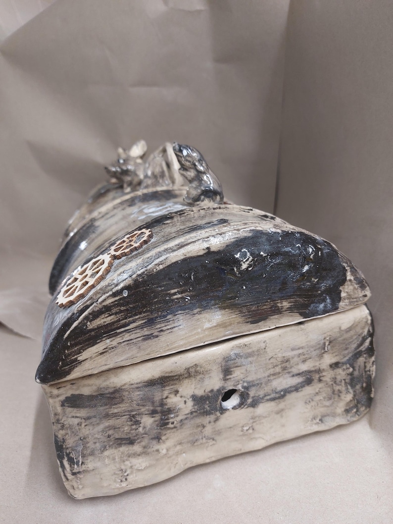 May include: A handmade ceramic box with a lid. The box is mainly off-white with black and grey accents, and a wood-grain texture. Decorative wooden gears are affixed to the side, and small animal figurines adorn the top. A small hole is visible on the front.