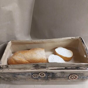 May include: A rectangular bread basket with a distressed, grey-washed finish. It contains a loaf of crusty bread and two slices. Decorative wooden cogs adorn the front of the basket, adding a unique touch.
