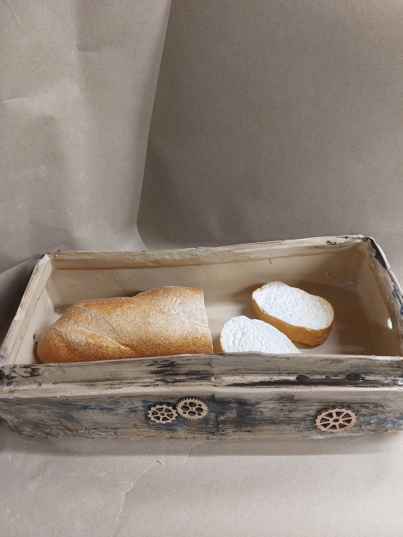 May include: A rectangular bread basket with a distressed, grey-washed finish. It contains a loaf of crusty bread and two slices. Decorative wooden cogs adorn the front of the basket, adding a unique touch.