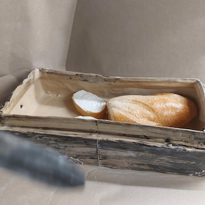 May include: A wooden bread bin with a loaf of bread and a slice inside. The bread has a golden-brown crust and a light interior. The bin is made of weathered wood with metal accents.