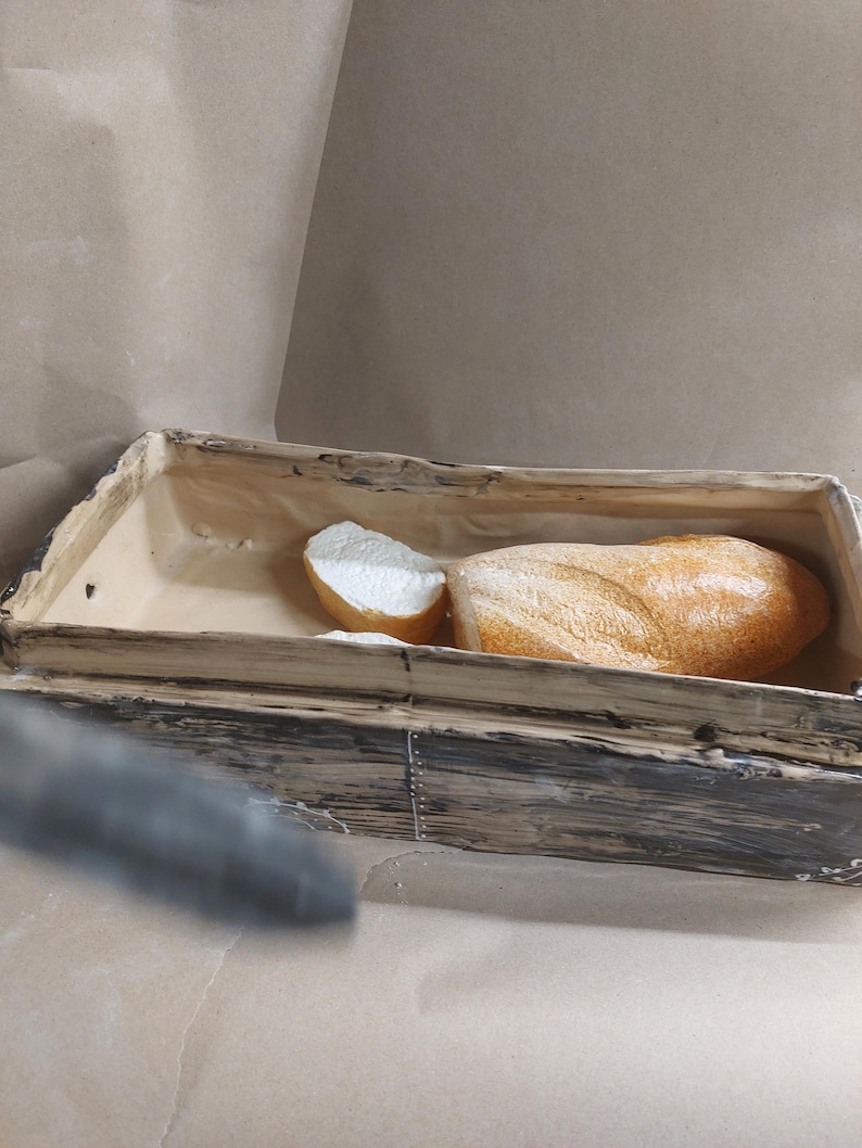 May include: A wooden bread bin with a loaf of bread and a slice inside. The bread has a golden-brown crust and a light interior. The bin is made of weathered wood with metal accents.