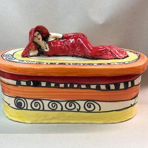 Può includere: Scatola in ceramica dipinta a mano con una figura reclinata sul coperchio. La scatola presenta una vivace combinazione di colori rosso, arancione, giallo e nero. La figura indossa un abito e un cappello rossi.