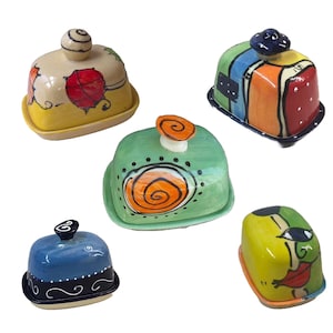 Könnte beinhalten: Fünf Keramik-Butterdosen mit farbenfrohen handbemalten Designs. Die Dosen sind in verschiedenen Farben, darunter Gelb, Grün, Blau, Rot und Orange. Jede Dose hat ein einzigartiges Design, darunter Spiralen, Punkte und abstrakte Formen.