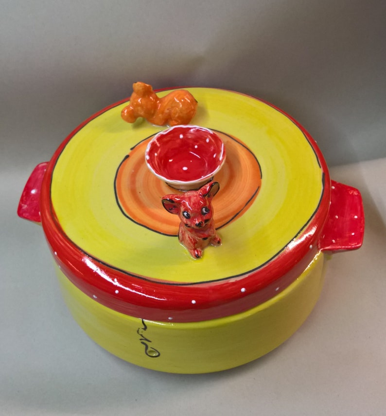 May include: Yellow ceramic container with a red lid and white polka dots. The lid has a small red bowl and two small ceramic animal figurines, one orange and one red.