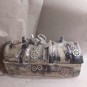 Peut inclure: Une boîte en céramique rectangulaire faite à la main avec une esthétique steampunk. La boîte est peinte dans des tons de gris et de marron, ornée d'engrenages en bois et d'une figurine de petite souris sur le dessus. Le couvercle est légèrement incurvé.