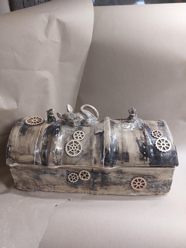 May include: A handcrafted, rectangular ceramic box with a steampunk aesthetic. The box is painted in shades of grey and brown, adorned with wooden cogs and a small mouse figurine on top. The lid is slightly curved.