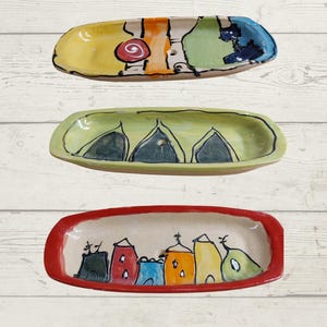 May include: Three hand-painted ceramic trays. The top tray is yellow, orange, green, and blue with abstract designs. The middle tray is light green with black outlines. The bottom tray has a red border and features colourful buildings.