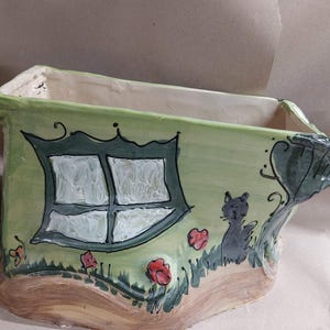 May include: A whimsical, hand-painted ceramic planter in shades of green, brown, and white. The planter features a house design with a window, a black cat, and red flowers. The overall design is charming and decorative.