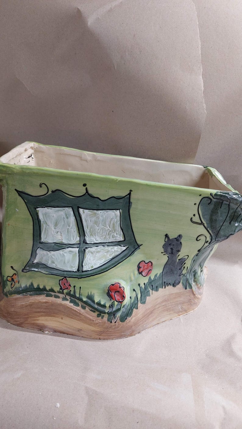 May include: A whimsical, hand-painted ceramic planter in shades of green, brown, and white. The planter features a house design with a window, a black cat, and red flowers. The overall design is charming and decorative.