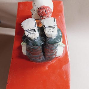 May include: A ceramic figurine of a seated figure with a white head, red hair, and dark green clothing. The figure is resting on a bright red, rectangular base. The figurine has a whimsical, folk art style.