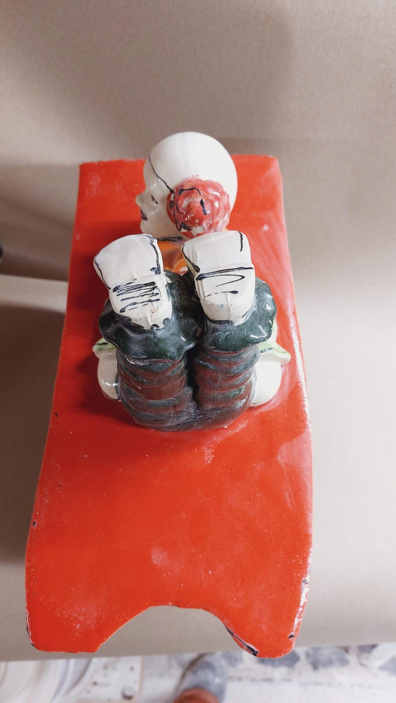 May include: A ceramic figurine of a seated figure with a white head, red hair, and dark green clothing. The figure is resting on a bright red, rectangular base. The figurine has a whimsical, folk art style.