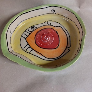 Könnte beinhalten: Handbemalte Keramikschale mit einzigartigem Design. Die Schale hat einen grünen Rand, einen beigefarbenen äußeren Bereich und ein lebendiges Inneres mit gelben, orangefarbenen und roten konzentrischen Kreisen. Schwarze Linien umreißen das Design. Ovale Form.