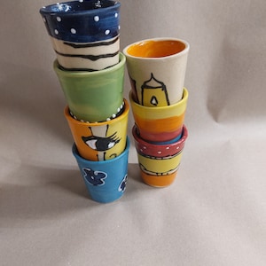 Könnte beinhalten: Ein Stapel von sechs bunten Keramikbechern mit verschiedenen handbemalten Designs. Die Becher sind pyramidenförmig gestapelt, wobei der oberste Becher blau mit weißen Punkten ist. Die anderen Becher zeigen Designs wie ein Auge, eine Blume und ein Gebäude.
