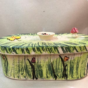 Peut inclure: Une boîte en céramique peinte à la main avec un couvercle représentant une scène de jardin fantaisiste. La boîte est peinte avec de l'herbe verte, des fleurs et un papillon jaune. Un petit bol et un champignon sont sur le couvercle.