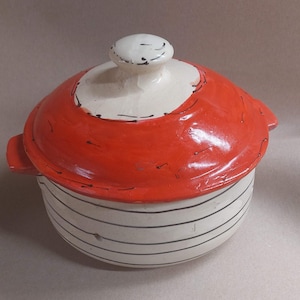 May include: A ceramic cooking pot with a lid. The pot is white with horizontal black stripes. The lid is red with a white rim and a white knob. The pot has two small handles on either side.