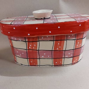 May include: Hand-painted ceramic bread box with a red and cream plaid pattern. The lid is red with white polka dots and a small, off-white ceramic knob. The box has a rectangular shape with rounded edges.