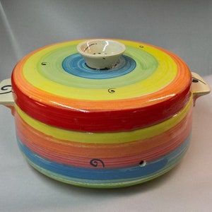 Può includere: Una pirofila rotonda in ceramica con coperchio, dipinta con strisce arcobaleno concentriche. Il coperchio ha un piccolo pomello bianco. La pirofila ha due manici color crema con disegni a spirale neri. Un oggetto da cucina vivace e colorato.