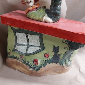 May include: A whimsical ceramic sculpture featuring a figure with a smiling face and a leg extending upwards. The figure is atop a house-shaped base with a red roof, a green wall, a window, and a black cat. Red flowers and green grass are painted on the base.