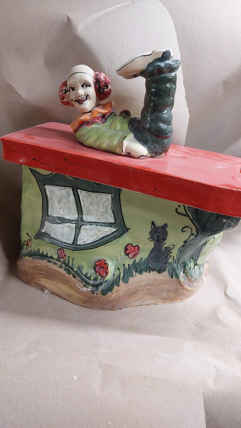 May include: A whimsical ceramic sculpture featuring a figure with a smiling face and a leg extending upwards. The figure is atop a house-shaped base with a red roof, a green wall, a window, and a black cat. Red flowers and green grass are painted on the base.