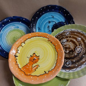 Könnte beinhalten: Eine Sammlung farbenfroher, handbemalter Keramikteller. Die Teller weisen verschiedene Designs auf, darunter eine Katze, Wirbel und Punkte. Farben sind Blau, Grün, Gelb, Orange und Braun. Die Teller sind leicht überlappend angeordnet.