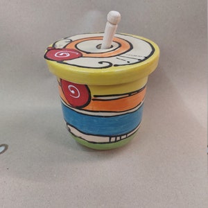 Può includere: Un vasetto di miele in ceramica giallo con un mestolo di legno. Il vasetto ha un design colorato con accenti arancioni, blu e rossi.