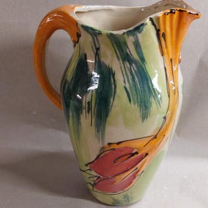 Könnte beinhalten: Eine handbemalte Keramikkrug mit einem lebendigen Blumendesign. Der Krug hat einen gebogenen Griff und einen Ausguss. Der Körper ist mit grünen und orangen Pinselstrichen und einem roten Blumendesign bemalt. Der Krug ist ein dekorativer Artikel.