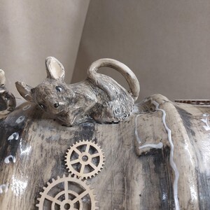 May include: A ceramic sculpture of a mouse figurine on a textured, cylindrical form. The mouse is grey with black details. Two wooden gear embellishments are attached to the surface, giving a steampunk look.