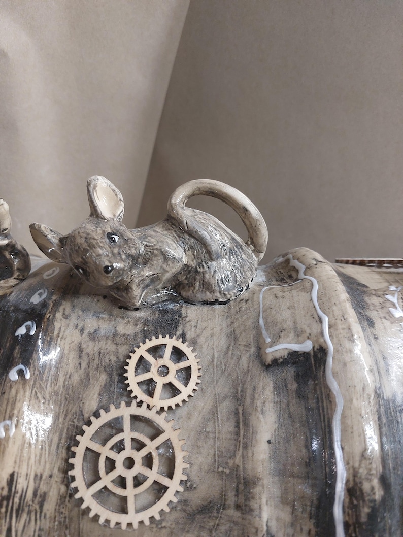 May include: A ceramic sculpture of a mouse figurine on a textured, cylindrical form. The mouse is grey with black details. Two wooden gear embellishments are attached to the surface, giving a steampunk look.