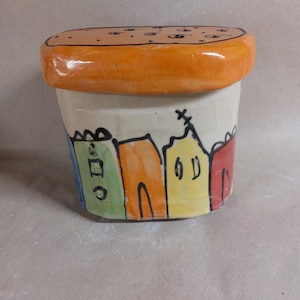 Può includere: Un contenitore in ceramica con coperchio arancione con puntini neri. Il contenitore bianco è decorato con una colorata scena di villaggio dipinta attorno alla base. La scena del villaggio comprende edifici blu, verdi, arancioni, gialli e rossi.
