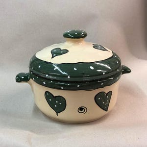 May include: A cream-colored ceramic casserole dish with a lid. The dish has a green and white polka dot design with green hearts. The lid has a knob on top.