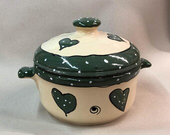 May include: A cream-colored ceramic casserole dish with a lid. The dish has a green and white polka dot design with green hearts. The lid has a knob on top.