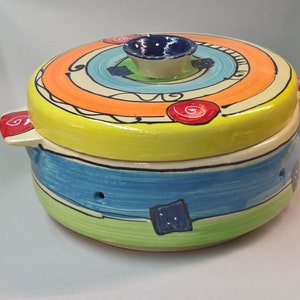 Könnte beinhalten: Ein farbenfroher Keramik-Auflauf mit Deckel. Der Deckel hat konzentrische Kreise in Orange, Blau und Gelb mit roten und schwarzen Akzenten. Der Sockel ist in Blau, Grün und Gelb bemalt. Er hat zwei kleine Griffe und einen kleinen Knopf auf dem Deckel.