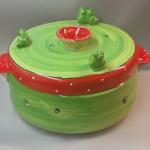 May include: A ceramic pot with a lid, painted in shades of green and red. The lid is green with a red band featuring white polka dots. Three small green frog figurines are on the lid. The pot has a small red bowl in the center.