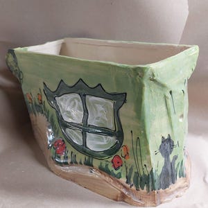 May include: Hand-painted ceramic planter in a whimsical house design. The rectangular planter features a green exterior with a window, flowers, and a cat illustration. The base is a light brown color.