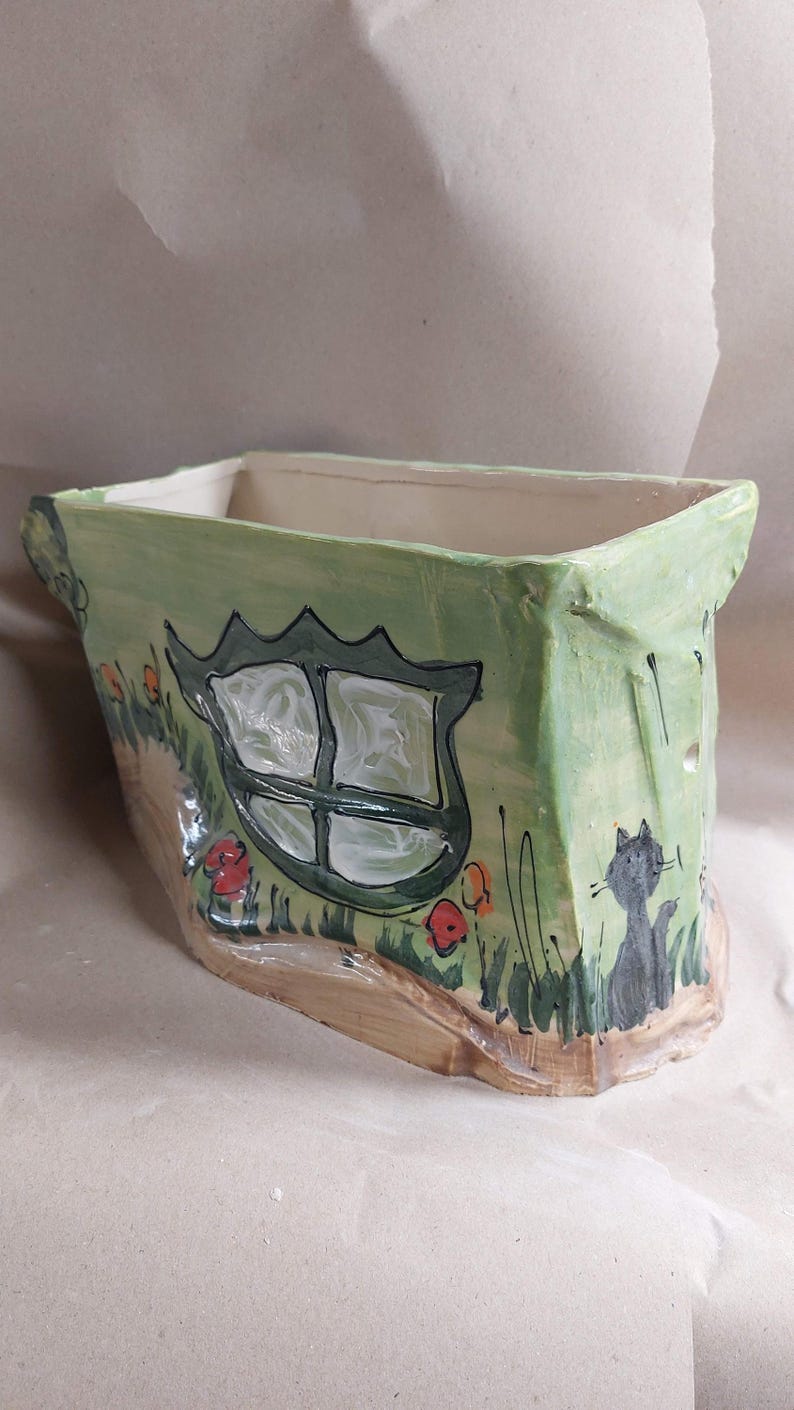 May include: Hand-painted ceramic planter in a whimsical house design. The rectangular planter features a green exterior with a window, flowers, and a cat illustration. The base is a light brown color.
