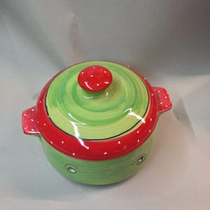 Könnte beinhalten: Ein runder Keramiktopf mit Deckel. Der Topf ist grün mit roten Akzenten und weißen Tupfen bemalt. Der Deckel hat einen roten Knopf mit weißen Tupfen. Der Topf hat zwei kleine Griffe. Ein dekorativer Küchenartikel.