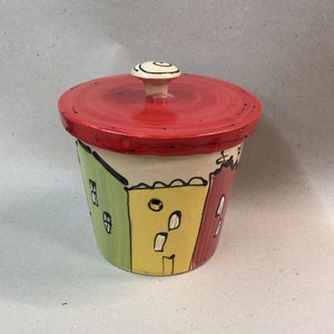 Peut inclure: Un pot en céramique peint à la main avec un couvercle rouge et un bouton blanc. Le corps du pot est décoré de formes colorées en forme de maison en vert, jaune et rouge, soulignées en noir. Le couvercle est rouge avec un contour noir.